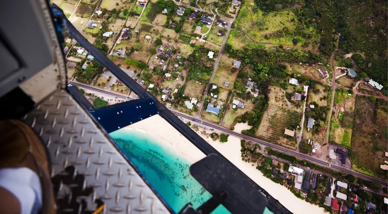 Straight-down view past a helicopter skid to a reef and beach, taken on a doors-off helicopter tour in Hawaii