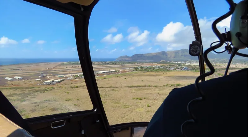 View forward through a helicopter windscreen on approach to a Hawaiian airfield, instruments visible in frame