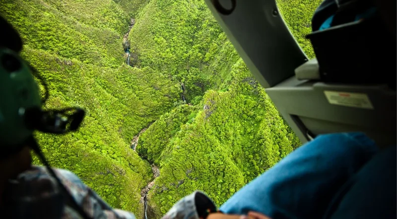 View from inside a helicopter cabin over a deep green valley threaded with waterfalls, passengers in headsets in frame