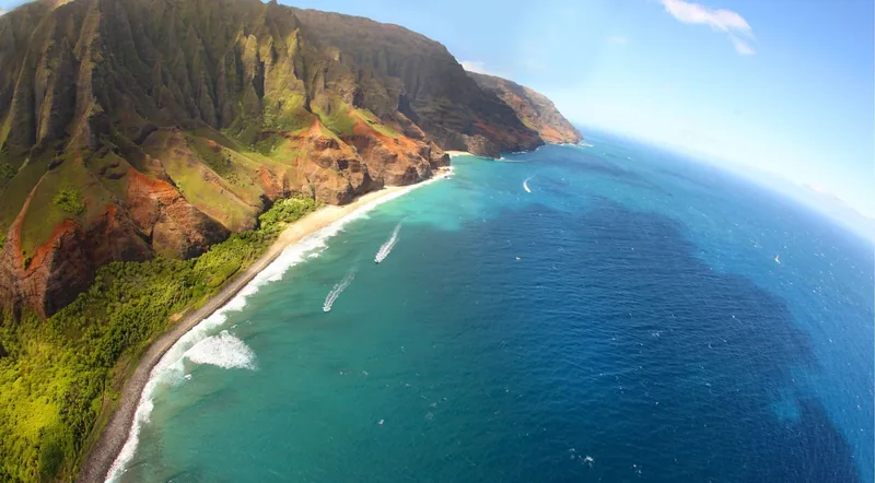 Wide aerial view along the Nā Pali coastline with small boats far below on the blue Pacific