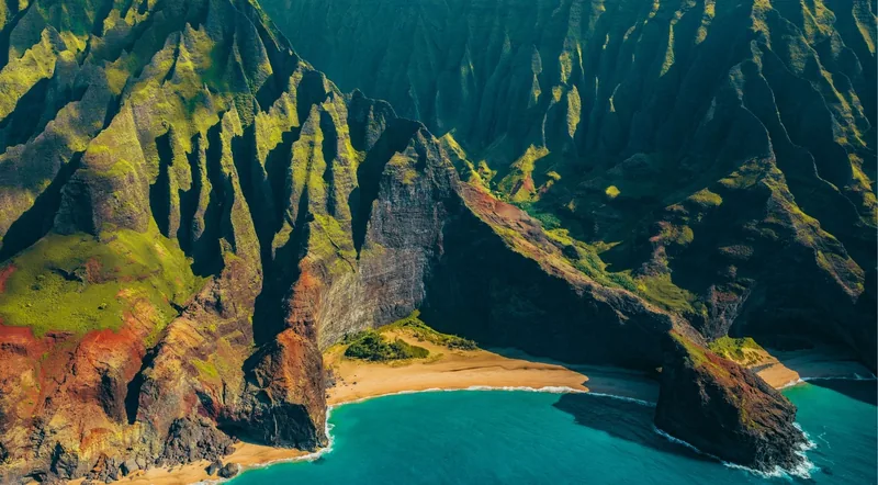 Red and green sea cliffs meeting a curved sand beach and turquoise water on Kauai's Nā Pali Coast